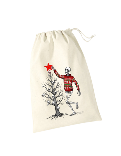 beiges Geschenksackerl aus Baumwolle mit Zugkordel mit weihnachtlichem Design: Skelett im Weihnachtspulli schmückt kahlen Weihnachtsbaum mit Totenköpfen und Knochen, steckt gerade roten Stern oben drauf